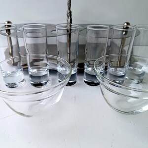 May include: A clear glass shot glass set with a silver metal serving tray. The set includes shot glasses and small glass bowls. The shot glasses are tall and slender, while the bowls are round and shallow. The set is designed for serving drinks.