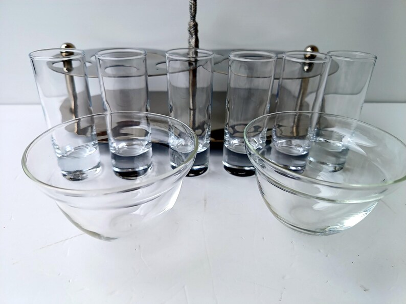 May include: A clear glass shot glass set with a silver metal serving tray. The set includes shot glasses and small glass bowls. The shot glasses are tall and slender, while the bowls are round and shallow. The set is designed for serving drinks.