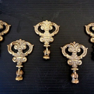 May include: Five antique brass key pulls with ornate, symmetrical designs. Each pull features a decorative top with scrolling details and a central design element. The pulls are mounted on a dark surface, showcasing their aged, golden color.
