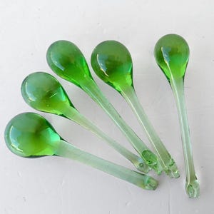 Puede incluir: Cinco cucharas de vidrio verde con mangos largos y delgados y cuencos redondeados están dispuestas en diagonal. Las cucharas tienen un color verde vibrante y transparente, y los mangos son de un tono más claro. Las cucharas son probablemente decorativas o para servir.