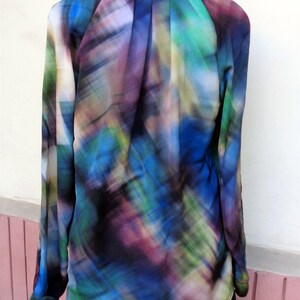 May include: A long-sleeved blouse with a vibrant, abstract pattern in shades of blue, green, purple, and black. The blouse has a collared neckline and is partially tucked into a black garment. The fabric appears to be lightweight and flowing.