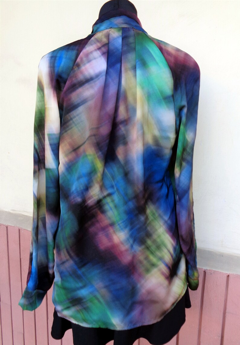 May include: A long-sleeved blouse with a vibrant, abstract pattern in shades of blue, green, purple, and black. The blouse has a collared neckline and is partially tucked into a black garment. The fabric appears to be lightweight and flowing.