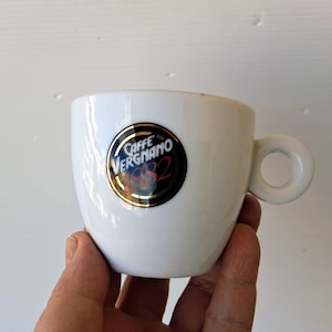 Könnte beinhalten: Eine weiße Espressotasse aus Keramik mit einem abgerundeten Henkel. Die Tasse hat ein rundes Logo mit den Worten "Caffe Vergnano 1882" in Gold und Schwarz. Die Tasse wird in einer Hand gehalten.