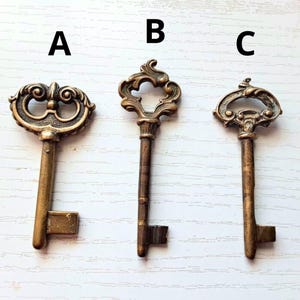 Pode incluir: Três chaves antigas de latão com desenhos ornamentados. A chave A tem um topo de flor-de-lis, a chave B tem um design em forma de coração e a chave C tem um topo curvo. Cada chave tem uma haste longa e uma palheta retangular.