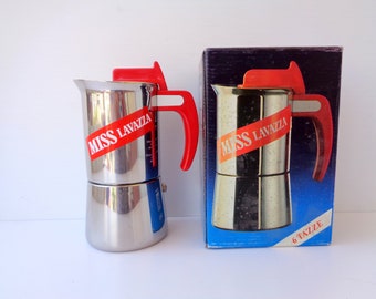 May include: A stainless steel Miss Lavazza stovetop espresso maker with a red handle and lid. The coffee maker is next to its original box, which also features the Miss Lavazza logo and the text "6 tazze".