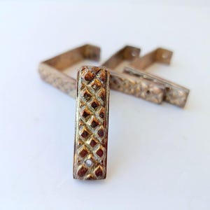 May include: A set of four rectangular metal drawer pulls with a textured diamond pattern. Each pull features a metallic gold and bronze finish with a single circular screw hole at the bottom. The hardware has an L-shaped profile for mounting.