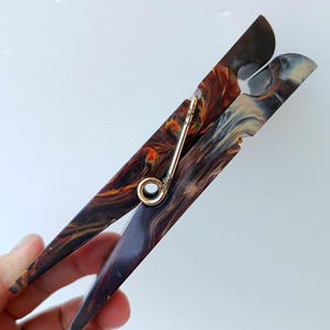 May include: A large clothespin featuring a marbled pattern in shades of dark brown, black, and cream with orange highlights. The two main pieces are connected by a central gold-toned metal spring mechanism that allows the clip to open and close.