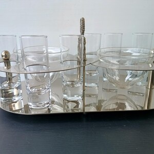 May include: A silver-toned serving tray with shot glasses and small bowls. The tray has a handle and a decorative design. The shot glasses and bowls are clear glass. The tray is on a dark surface.