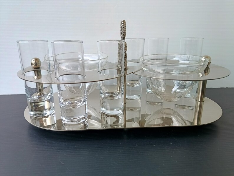 May include: A silver-toned serving tray with shot glasses and small bowls. The tray has a handle and a decorative design. The shot glasses and bowls are clear glass. The tray is on a dark surface.