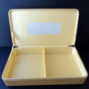 Puede incluir: Maleta rectangular abierta, de color amarillo pálido, con interior dividido. La maleta tiene un espejo rectangular y un ribete dorado. La maleta está abierta, revelando el interior, sobre un fondo oscuro.