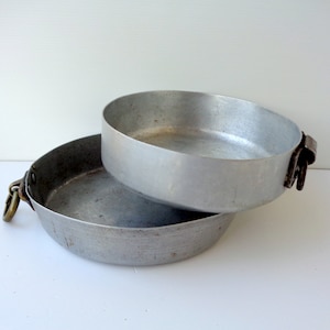May include: Two vintage aluminium pans. One pan is nested inside the other. The bottom pan has a handle with a brass ring. The top pan has two metal handles. The pans are silver and show signs of wear.