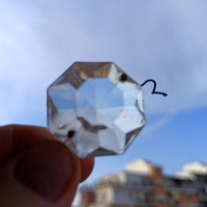 Op de afbeelding: Een helder, gefacetteerd, achthoekig glazen kristalprisma met een klein metalen lusje aan de bovenrand. Het transparante object heeft meerdere geometrische oppervlakken die het licht reflecteren en een doorkijk door het midden bieden.