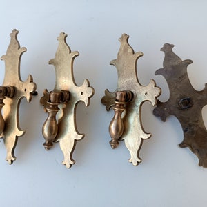 Könnte beinhalten: Vier antike Schubladengriffe aus Messing. Jeder Griff hat ein verziertes, stilisiertes Design mit einem dekorativen Griff. Die Griffe haben eine warme, gealterte Messingfarbe, was auf Vintage- oder geborgene Hardware hindeutet. Das Design ist symmetrisch und detailliert.