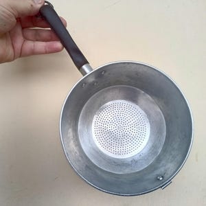 May include: A small, round, aluminium saucepan with a dark brown handle. The interior features a perforated metal insert. The pan is held by a hand against a light-coloured background. The handle is approximately 13 cm long.