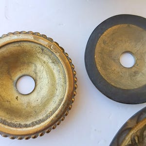 May include: Three circular metal components with central holes. One piece features a textured, beaded edge with a bright brass finish. A second piece is a flat, dark-toned metal disc. A third partial piece shows a decorative, ornate brass border.