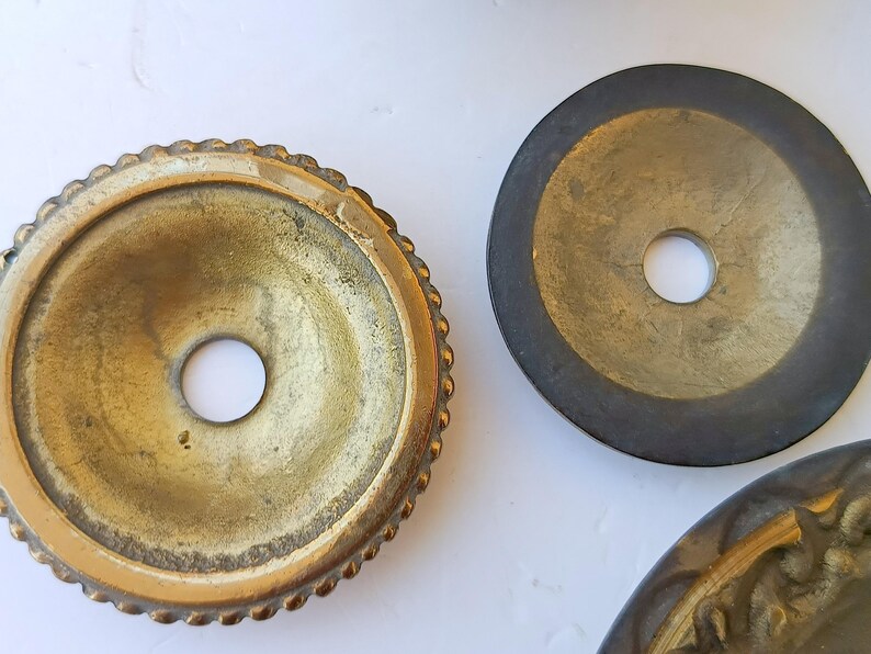 May include: Three circular metal components with central holes. One piece features a textured, beaded edge with a bright brass finish. A second piece is a flat, dark-toned metal disc. A third partial piece shows a decorative, ornate brass border.