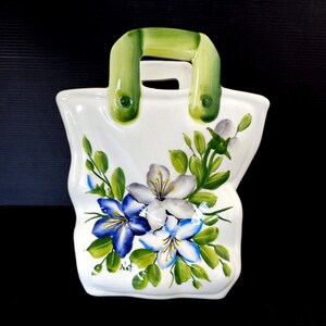 Può includere: Oggetto decorativo in ceramica bianca a forma di borsa, con manico verde. La borsa è decorata con fiori blu e bianchi dipinti a mano e foglie verdi. La base della borsa è leggermente curva, e il manico è di colore verde.
