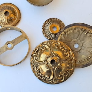 May include: A collection of brass hardware components including a circular mounting bracket with a central crossbar, two small decorative rosettes with scalloped edges, and two larger circular plates featuring intricate embossed scrollwork patterns.
