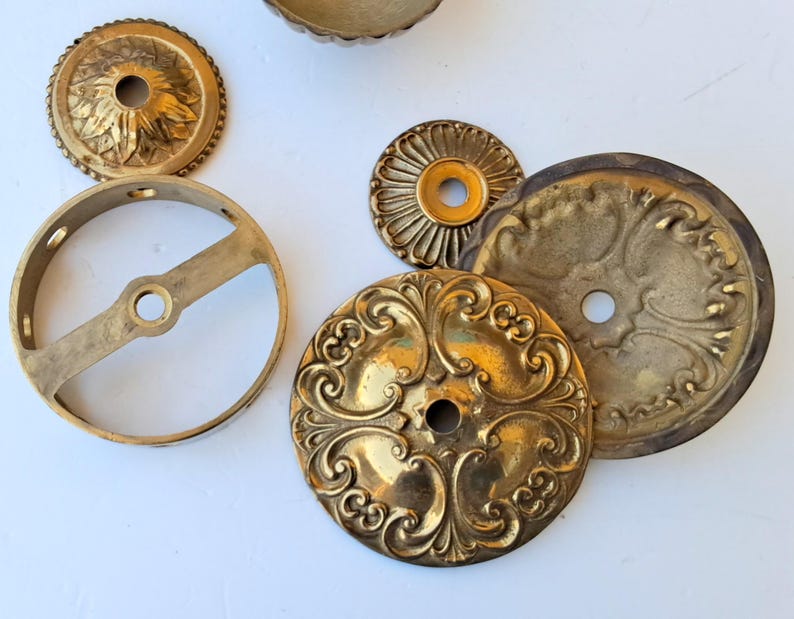 May include: A collection of brass hardware components including a circular mounting bracket with a central crossbar, two small decorative rosettes with scalloped edges, and two larger circular plates featuring intricate embossed scrollwork patterns.