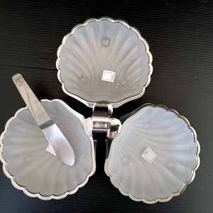 May include: A silver-toned, three-section serving dish with a seashell design. Each section is frosted glass with a scalloped silver rim. A matching silver butter knife rests on one section. The dish is designed to swivel.