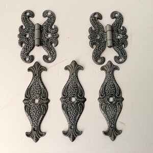 May include: Six decorative, ornate metal hinges and door pulls. The hinges have a butterfly design, while the pulls are elongated with a textured surface. The hardware is painted in a distressed gray finish, suitable for a rustic or vintage aesthetic.