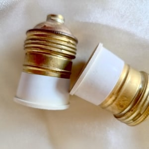 Może przedstawiać: Dwa zabytkowe oprawki żarówek. Oprawki mają mosiężne wykończenie z białą podstawą. Oprawki są w pozycji poziomej na miękkim, kremowym tle. Jedna oprawka jest częściowo zdemontowana.