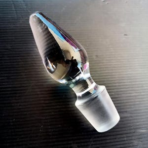 May include: A clear glass bottle stopper with a conical shape and a frosted base. The stopper is designed to fit into a bottle and has a smooth, reflective surface. The glass reflects light, creating a play of colors.