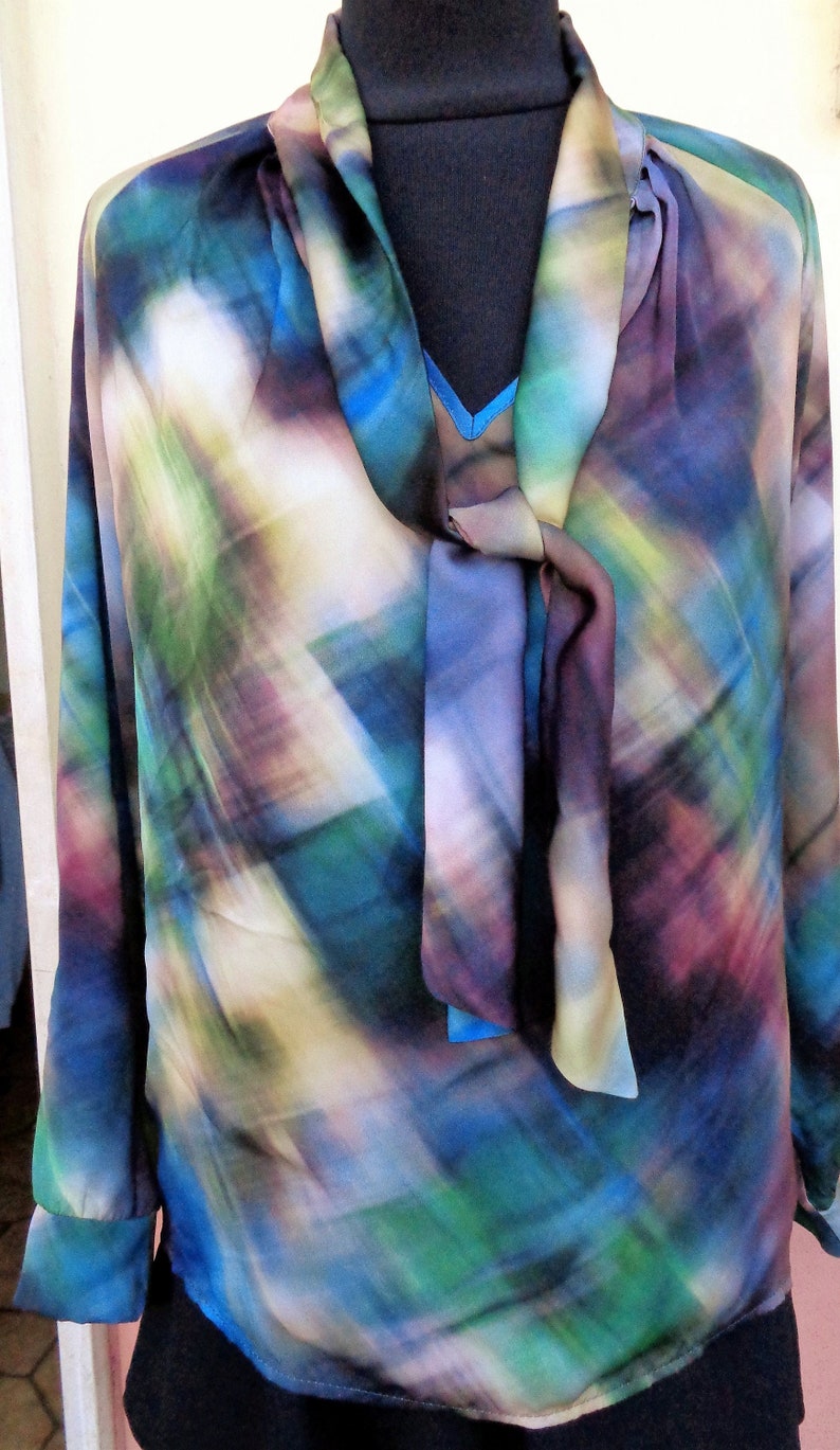 May include: A long-sleeved blouse with a colorful abstract pattern in shades of blue, green, purple, and white. The blouse features a tie-neck detail and buttoned cuffs. The garment is displayed on a black mannequin.