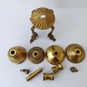 May include: An assortment of antique brass lamp parts, including decorative bases, caps, and connecting tubes. The components vary in size and shape, with a gold finish and ornate detailing. The parts are arranged on a white surface, showcasing their vintage aesthetic.