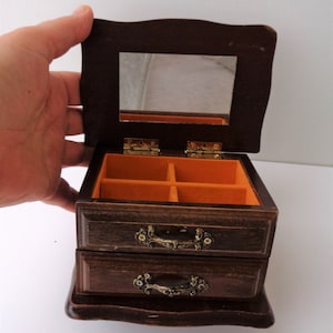 May include: A vintage wooden jewelry box with two drawers and a mirrored lid. The box is dark brown with ornate brass handles and hinges. The interior is lined with orange felt, and the box is open, revealing the compartments.