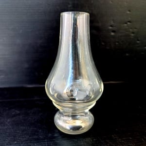May include: A clear glass lamp chimney with a bulbous base and a flared top. The glass has a slight pearlescent sheen and is designed to fit over a light source. The chimney is approximately 5 inches tall.