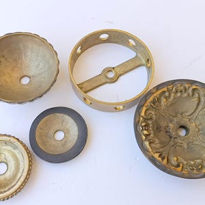May include: A collection of five brass hardware components including a scalloped bowl, a circular ring with a central crossbar, a flat disc with a dark rim, a small circular plate with a beaded edge, and a decorative plate with ornate relief.