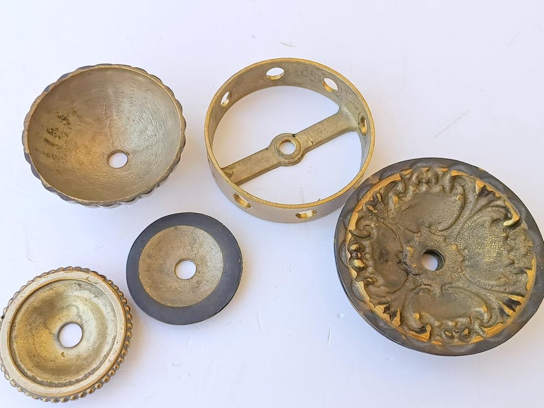 May include: A collection of five brass hardware components including a scalloped bowl, a circular ring with a central crossbar, a flat disc with a dark rim, a small circular plate with a beaded edge, and a decorative plate with ornate relief.