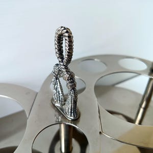 May include: Silver-toned metal serving tray with a decorative handle and tassel accents. The tray features multiple circular cutouts and a central vertical support, suggesting a serving or display function.