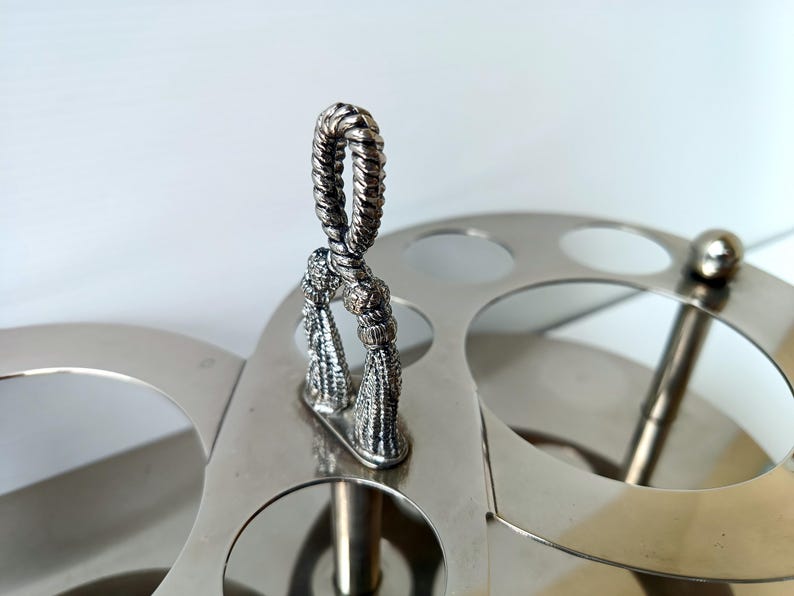 May include: Silver-toned metal serving tray with a decorative handle and tassel accents. The tray features multiple circular cutouts and a central vertical support, suggesting a serving or display function.