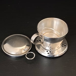 Könnte beinhalten: Ein vietnamesischer Kaffeefilter aus Edelstahl, auch Phin genannt, mit Deckel und Griff. Der Filter hat einen perforierten Boden und einen Griff für eine einfache Handhabung. Der Deckel hat einen kleinen Ring zum Anheben. Das Set ist zum Brühen von Kaffee konzipiert.