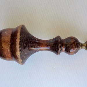 Puede incluir: Un remate de madera marrón oscuro con un anillo de latón en el extremo. El remate tiene una parte superior bulbosa y un cuerpo delgado y texturizado. La veta de la madera es visible y el acabado es brillante.