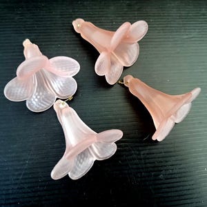 May include: Four pink plastic flower-shaped embellishments with a gold wire loop at the top. The flowers have five petals and a bell-shaped center.