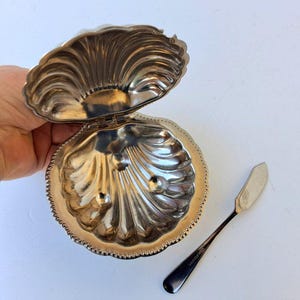 Pode incluir: Uma caixa de bugigangas prateada em forma de concha com uma tampa aberta, revelando um interior com três pequenas protuberâncias. Uma pequena faca de manteiga prateada repousa ao lado da caixa. A caixa e a faca estão sobre uma superfície branca.