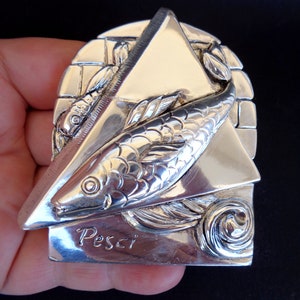 May include: A silver-toned metal zodiac charm featuring a stylised fish design with the word "Pesci" inscribed below. The charm is shaped like a triangle with a textured brick pattern on the back.