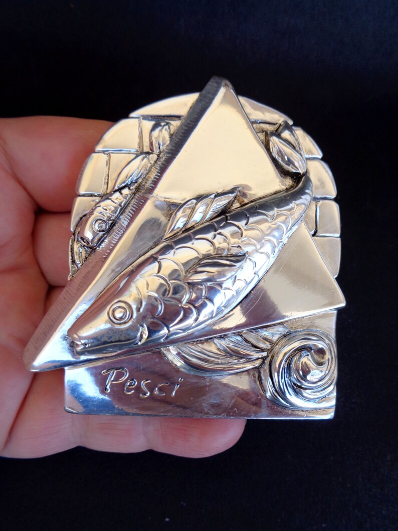 May include: A silver-toned metal zodiac charm featuring a stylised fish design with the word "Pesci" inscribed below. The charm is shaped like a triangle with a textured brick pattern on the back.