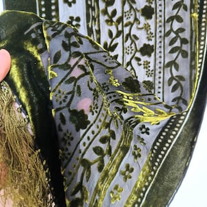 May include: Close-up of a dark green velvet scarf with a floral pattern and fringe. The scarf has a sheer, light-coloured background with dark green floral designs and a velvet border. The fringe is a matching green.