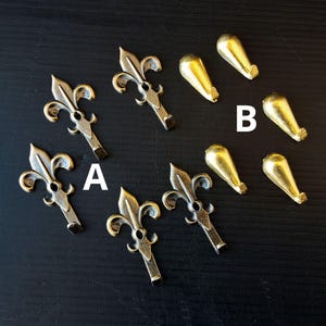 May include: A selection of antique brass-toned metal hardware, featuring fleur-de-lis shaped pulls and teardrop-shaped hooks. The hardware is displayed on a dark surface, with labels "A" and "B" denoting different styles. The fleur-de-lis pulls have an intricate, decorative design.