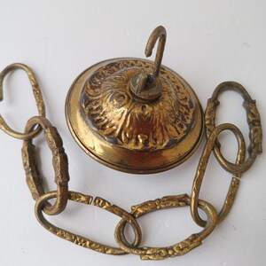Könnte beinhalten: Antike Deckenleuchtenkomponenten aus Messing, darunter ein dekoratives Baldachin mit Haken und eine Kette. Das Baldachin hat ein verziertes Design. Die Kette besteht aus ineinandergreifenden Gliedern. Die Komponenten sind in einem warmen Goldton gehalten.