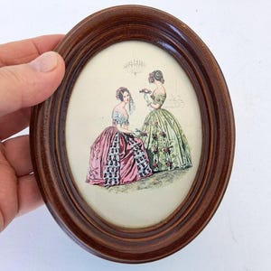 Può includere: Stampa ovale con cornice in legno raffigurante due donne in abiti elaborati. La stampa ha uno sfondo color crema con un lampadario e dettagli architettonici. Gli abiti sono rosa e verdi con accenti floreali.
