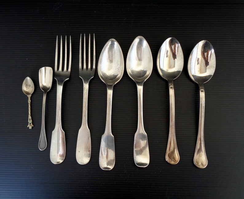 May include: A set of seven silver utensils, including a small spoon, a larger spoon, two forks, and three soup spoons. The utensils are arranged in a row on a black surface.