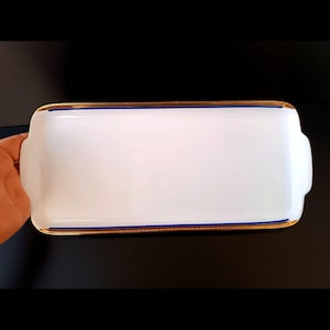 May include: A rectangular white serving tray with gold trim and blue accents. The tray has rounded edges and small handles on each end. The gold trim is a thin band that runs along the edge of the tray.