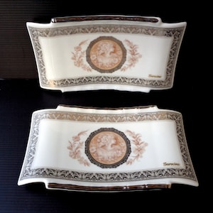Peut inclure: Deux plats de service en céramique blanche avec un motif décoratif. Chaque plat présente un médaillon ovale central avec une figure classique, entouré de motifs floraux et de volutes marron et or. Le mot "Saracino" est imprimé sur chaque plat.