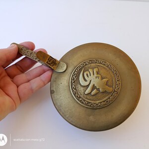 May include: A round, antique brass ashtray with a decorative handle and a central medallion featuring an embossed design. The ashtray has a circular shape and a tarnished finish, suggesting age and use. The handle is intricately detailed.