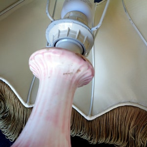 Op de afbeelding: Een roze keramische lampenvoet met een witte lampenkap van stof met een gouden franje. De lampenkap is bevestigd aan een metalen frame.
